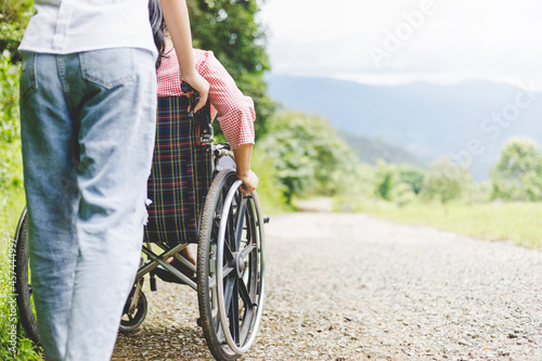 Disabled handicapped woman in wheelchair and care helper walking on mountain meadow park in sunny day. International Disability Day concept.