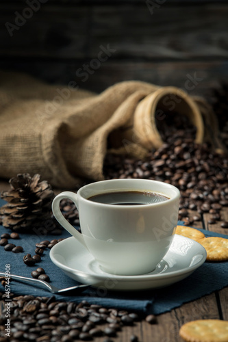 Hot coffee in a white coffee cup and many coffee beans placed around and sugar on a wooden table in a warm, light atmosphere, on dark background.
