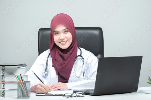 The Asian Muslim woman doctor was sitting at the patient's table and looked at the camera with a smiling and welcoming face.