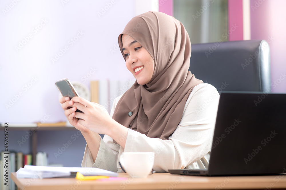 The office worker, an Asian Muslim woman, is sitting in front of her laptop computer at her desk and playing mobile phones and smiling happily at the office. The office worker, an Asian Muslim woman, is sitting in front of her laptop computer at her desk and playing mobile phones and smiling happily at the office.