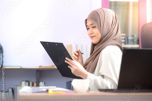 The office worker, an Asian Muslim woman, is sitting in front of the laptop computer at the desk and is looking at the files with success at the office.
