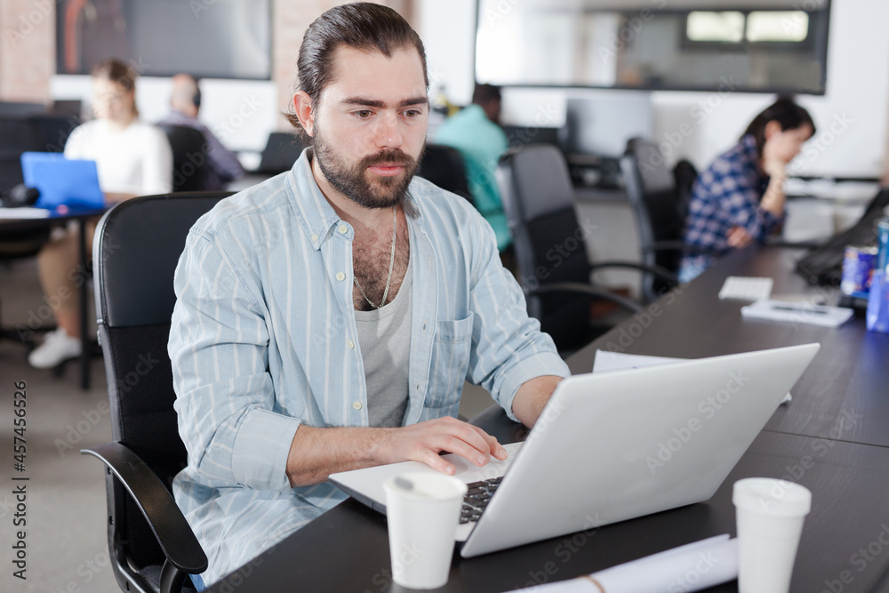 Portrait of bearded young man working on laptop in busy modern coworking office
