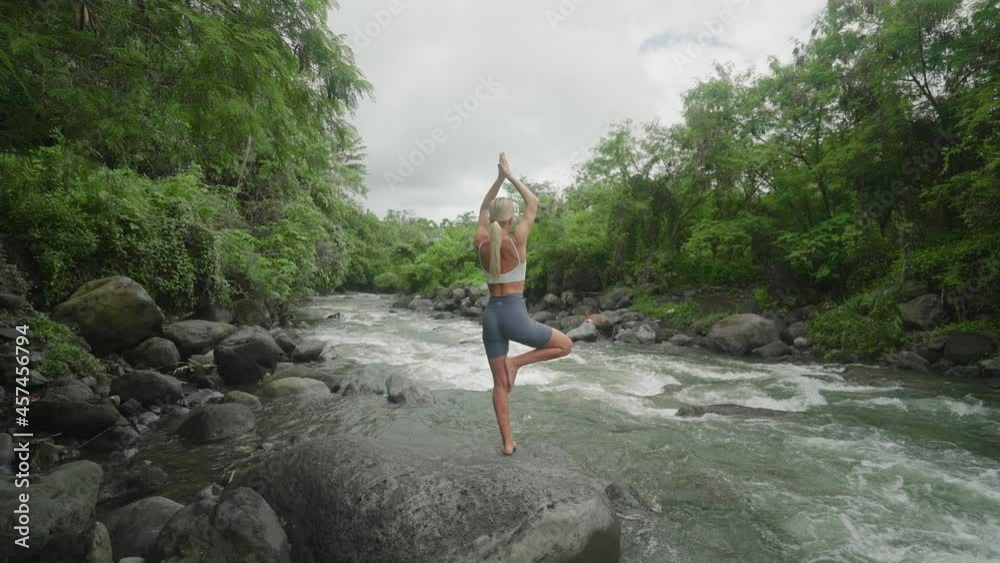 Behind blond woman practicing yoga beside tropical river, Tree Pose, Bali Behind blond woman practicing yoga beside tropical river, Tree Pose, Bali
