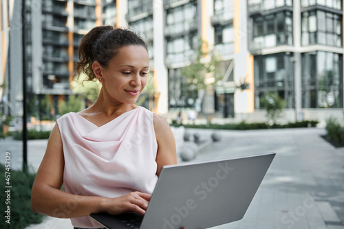 Attractive Hispanic young woman working on laptop, cutely smiling with toothy smile while looking at the screen monitor, standing on the urban buildings background. Business and communication concept © Taras Grebinets