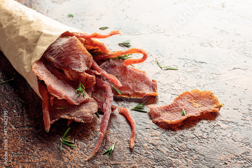 Slices of spicy dry-cured meat with rosemary in a paper bag on an old brown background.