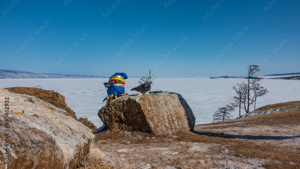 Picturesque boulders lie on the ground, on the shore of a frozen lake. A pigeon is sitting on a rock. Nearby, a pillar is visible, tied with ritual multicolored ribbons. Baikal. Picturesque boulders lie on the ground, on the shore of a frozen lake. A pigeon is sitting on a rock. Nearby, a pillar is visible, tied with ritual multicolored ribbons. Baikal.
