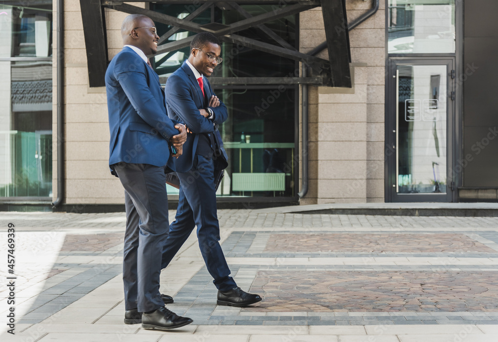 Two dark-skinned african american businessmen in suits and glasses walk outdoors in the city and discuss business