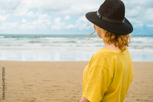 closeup of unrecognizable woman in yellow shirt and black hat standing on the beach looking at the sea