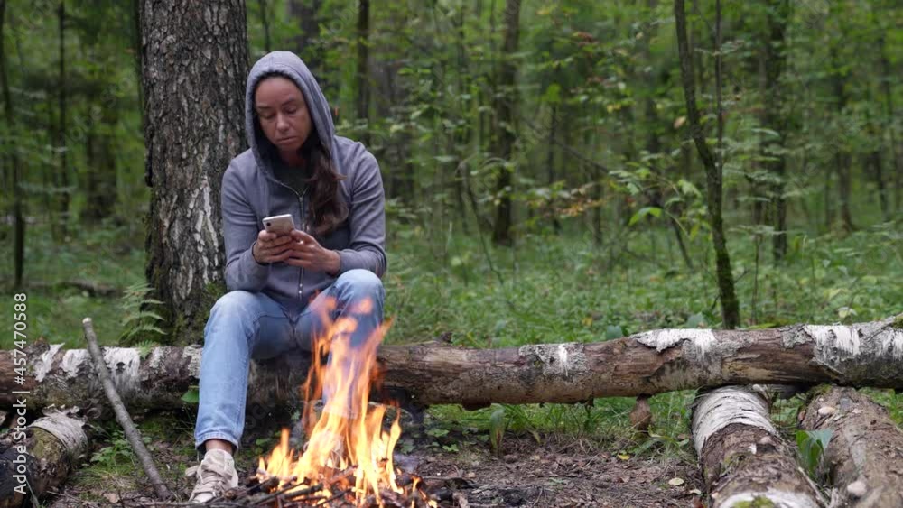An adult female tourist in a hoodie is sitting in the forest by the fire with a mobile phone in her hands for a social network An adult female tourist in a hoodie is sitting in the forest by the fire with a mobile phone in her hands for a social network