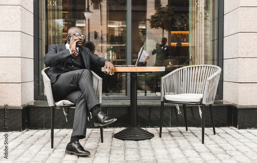 Portrait of a black African American businessman in a suit sitting in a city cafe outdoors and talking on the phone.