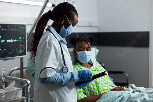 Medic of african ethnicity helping sick patient at clinic in hospital ward. Doctor using medical equipment and technology for young woman healthcare and treatment. Black person in bed