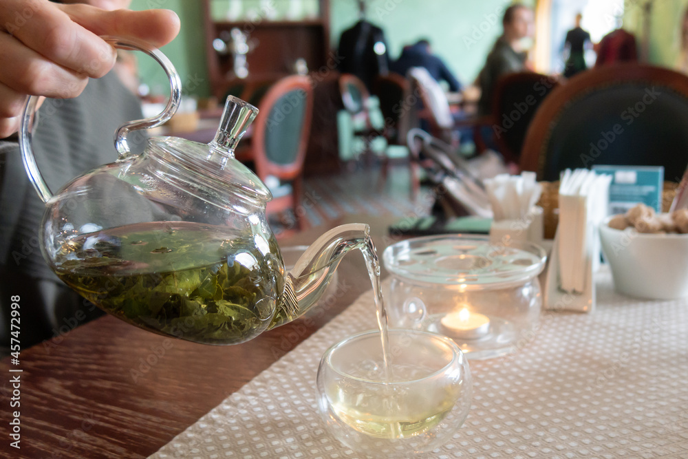 Woman pouring green tea from glass teapot into cups in cafe with blurred background