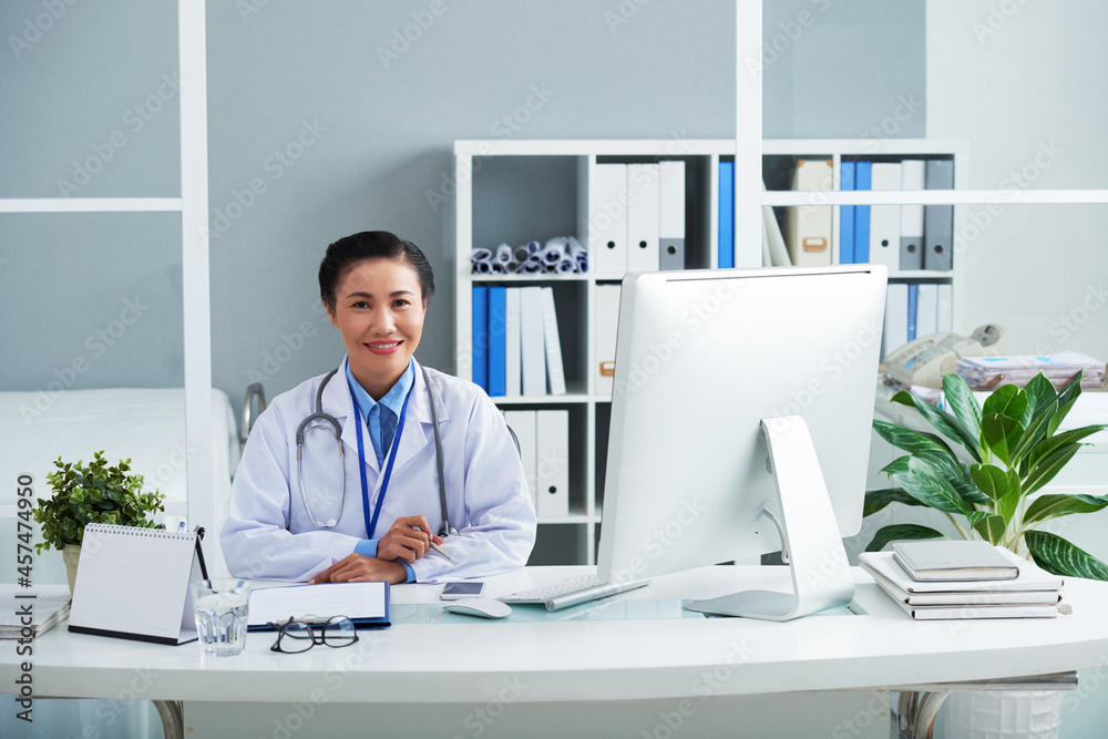 Beautiful smiling young Vietnamese chief physician signing documents when working at her office desk in clinic Beautiful smiling young Vietnamese chief physician signing documents when working at her office desk in clinic