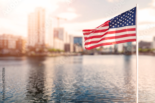 Waving flag of United States of America in focus. Modern town out of focus in the background. Warm sunny day. Sun flare. Abstract city concept photo