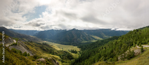 view from the top of the mountain pass to the valley drenched in the sun breaking through the clouds on a warm summer day  mountains  road 