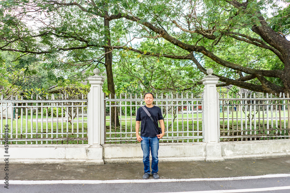 Asian Black T-Shirt is standing in front of white vintage fence beside the road in the afternoon time.
