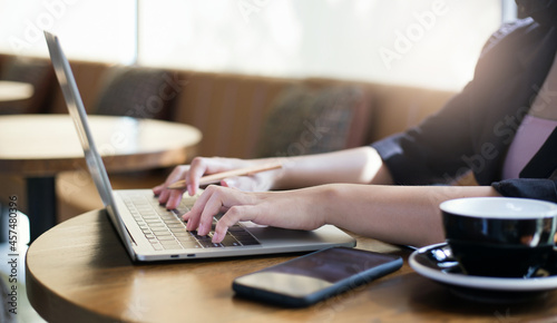 Focus on hand young asian businesswoman using laptop working and meeting online with mobile phone and coffee cup on wooden table
