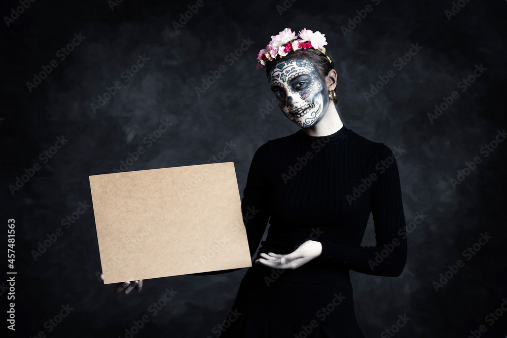 Woman with face painted as a Catrina and flowers in her hair on dark background pointing at poster. Day of the Dead - obrazy, fototapety, plakaty 