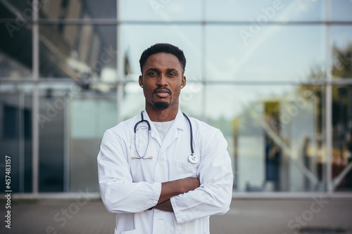 Portrait of a young serious afro american doctor, near the clinic, in a white medical gown looking at the camera © Liubomir