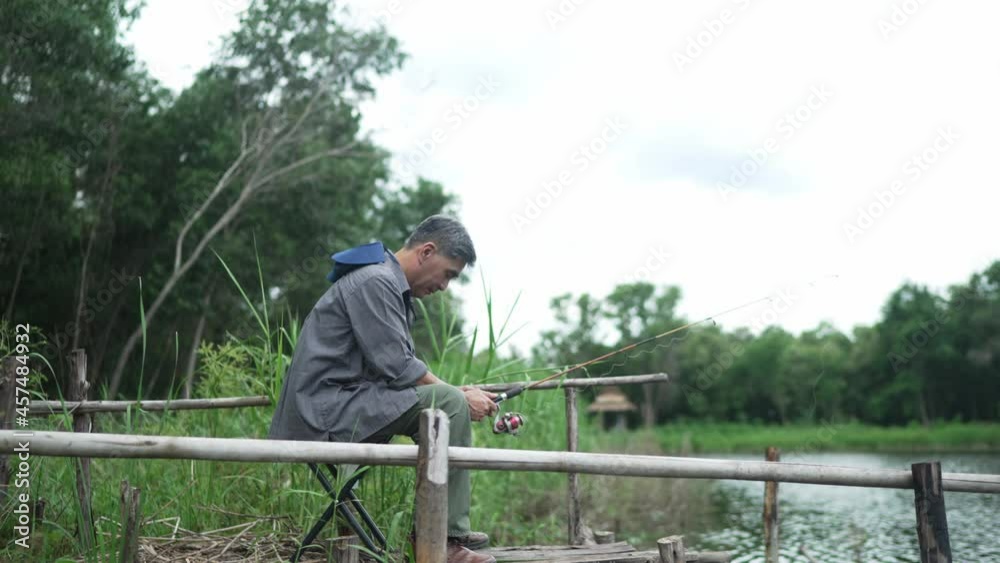 Elderly Asian man, gray hair, hat, holding a fishing rod, sits and sleeps fishing. On the edge of the lake, on vacation, retirement, hobbies for the elderly, concept Elderly Asian man, gray hair, hat, holding a fishing rod, sits and sleeps fishing. On the edge of the lake, on vacation, retirement, hobbies for the elderly, concept