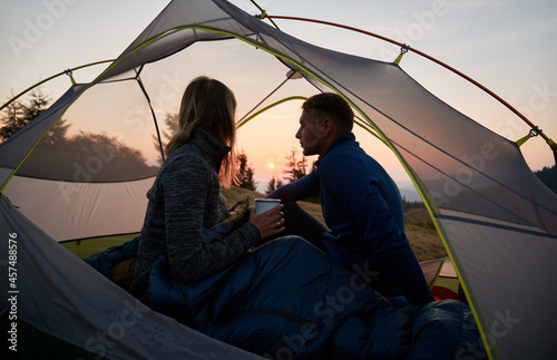 Girl sitting in tent, covered with sleeping bag, with her guy, holding metal cup and enjoying the view of sky during sunrise over the mountain hills. Concept of hiking, camping and relationships.