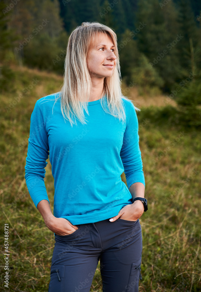 Young sporty blonde woman standing on green lawn and looking to the right on background of green forest. Lady watching into the distance with calm smile on her face outdoors during walk. Young sporty blonde woman standing on green lawn and looking to the right on background of green forest. Lady watching into the distance with calm smile on her face outdoors during walk.