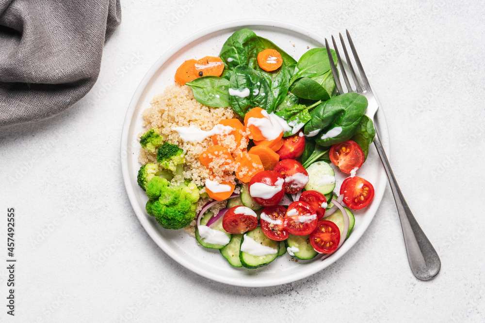 Salad with quinoa, spinach, broccoli, tomatoes, cucumbers, carrots and yoghurt dressing served on plate with white background and copy space