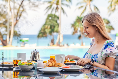 Young Beautiful Smiling Woman with Long Hair Eat Delicious Breakfast at Resort Restaurant by the Pool with Palms and Turquoise Sea on Background. Vacations or Holidays in Tropical Hotel © TravelMedia