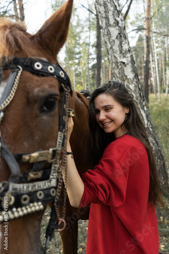 A beautiful long-haired girl in a red hoodie stands near the horse.