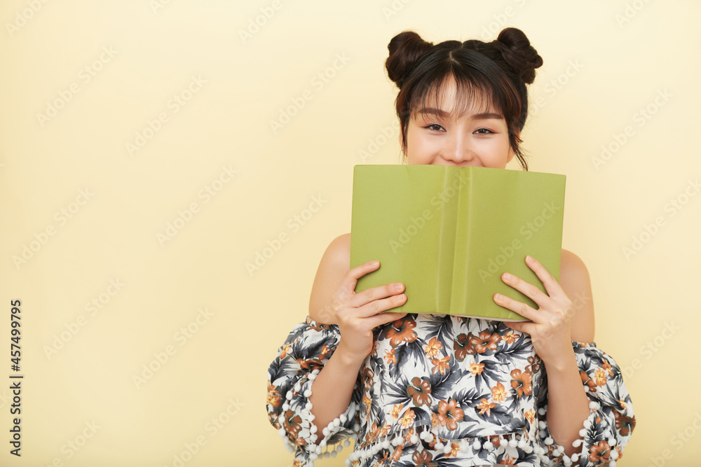 Happy pretty young Vietnamese woman hiding her smile behind opened book and looking at camera Happy pretty young Vietnamese woman hiding her smile behind opened book and looking at camera