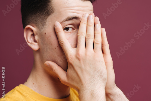 Handsome man with brown eyes wearing yellow t shirt peeking in shock covering face and eyes with hand, looking through fingers with embarrassed expression on crimson background. photo