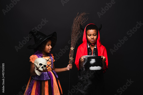african american children in halloween costumes holding skull and witch cauldron with potion isolated on black