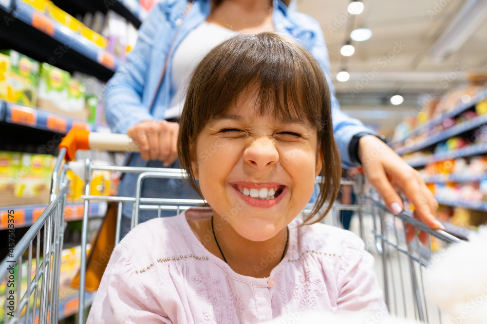 Close up photo of a smiling wide girl showing her teeth being in the grocery cart doing shopping together with her mother. Close up photo of a smiling wide girl showing her teeth being in the grocery cart doing shopping together with her mother.
