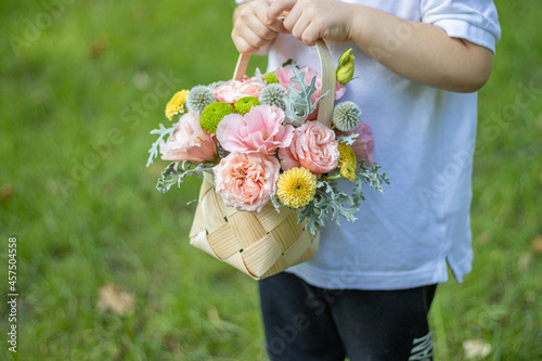 A little boy holds a basket of beautiful flowers in his hands. Happy kid is in the park and has a smiley face. Green blur background.