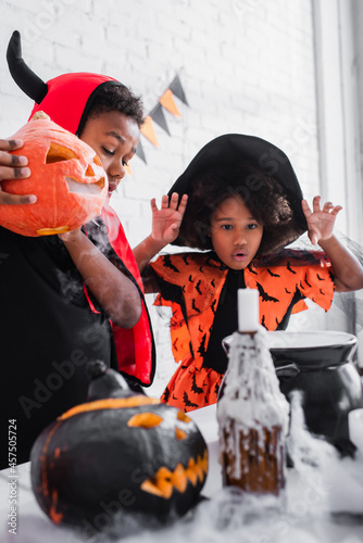 african american boy in halloween costume holding carved pumpkin while preparing potion near spooky sister in pointed hat