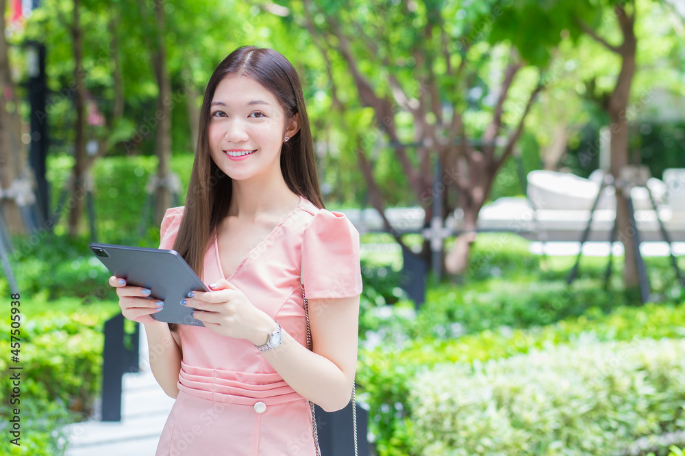 A professional Asian business woman with long hair is smiling in the garden while looking at the tablet in her hand, work from anywhere concept. A professional Asian business woman with long hair is smiling in the garden while looking at the tablet in her hand, work from anywhere concept.