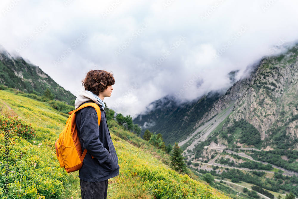 Portrait of a young teenager boy with curly hairs , with yellow backpack in a black jacket standing in the mountains in French Alps, looking away with pleasure. Hiking activities in summer.