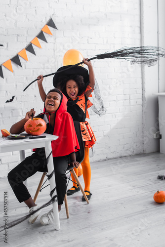 happy african american boy in halloween costume carving pumpkin near cheerful sister with broom