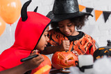african american boy in halloween costume looking at sister carving pumpkin