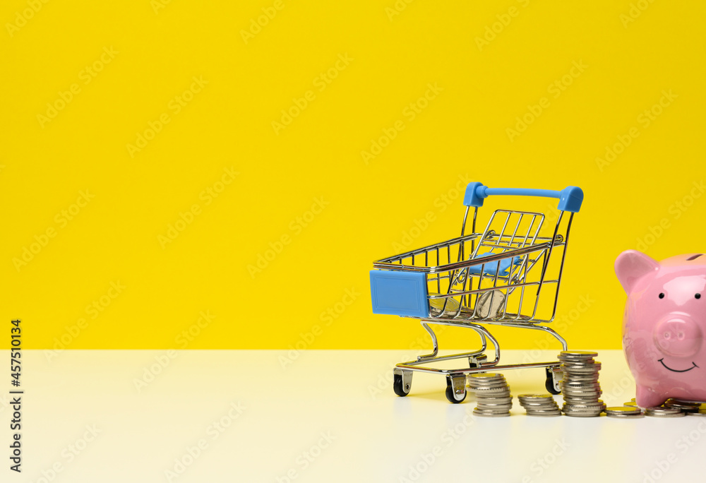metal miniature shopping cart with change and pink piggy bank on a white table, yellow background. Concept of saving budget, discounts