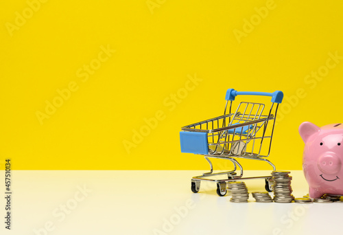 metal miniature shopping cart with change and pink piggy bank on a white table, yellow background. Concept of saving budget, discounts
