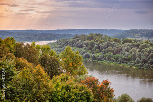 A river flows along the banks overgrown with trees and lush grass against the sunset sky. Rural landscape in autumn, city structures in the background.