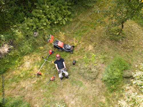 Gardener resting and sitting in a wheelbarrow tired after mowing the grass and cutting branches. Around it is a mower, a brushcutter and a chainsaw. View from above photo