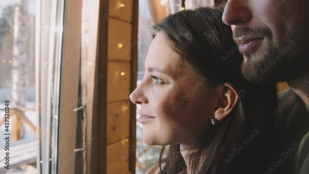 Handheld close up shot with slowmo of happy man and woman standing together by window and enjoying sunny winter day in their cottage