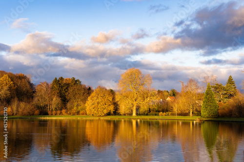 Small lake surrounded by forest with colorful trees at autumn cloudy day in Belgium. Panorama of a gorgeous park in autumn, a scenic landscape with pleasant warm sunshine. Trees reflections in water.