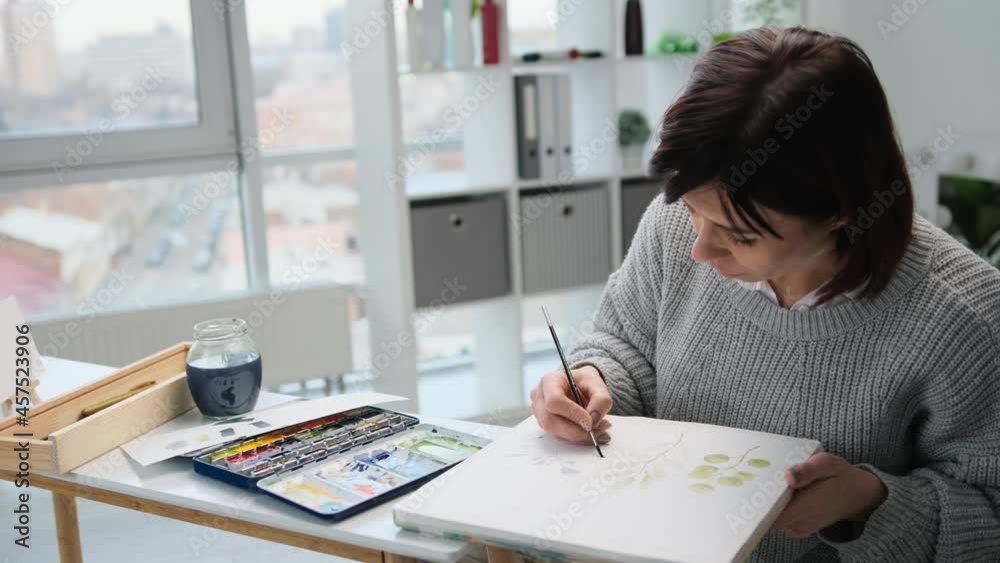 Woman painter sitting by the table and drawing with watercolor leaves on white paper in light room studio. Female person creates art work with aquarelle Woman painter sitting by the table and drawing with watercolor leaves on white paper in light room studio. Female person creates art work with aquarelle