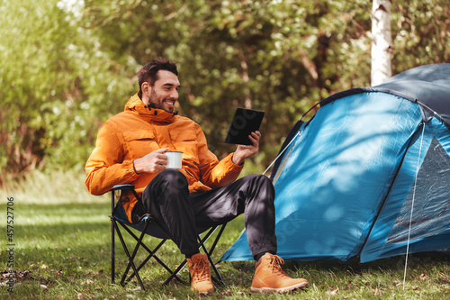 camping, tourism and travel concept - happy man with tablet pc computer drinking tea at tent camp photo