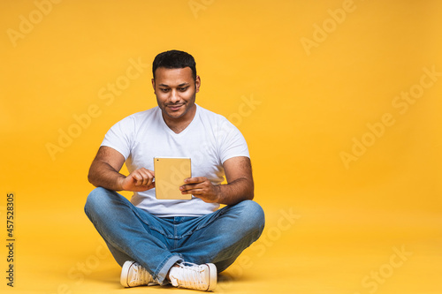 Portrait of young african american black indian man sitting on the floor using a tablet pc, isolated over yellow background. Ready for your design.