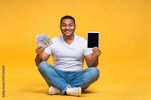 Portrait of african american indian black young man sitting on the floor holding dollar banknotes isolated over yellow background. Using tablet computer.