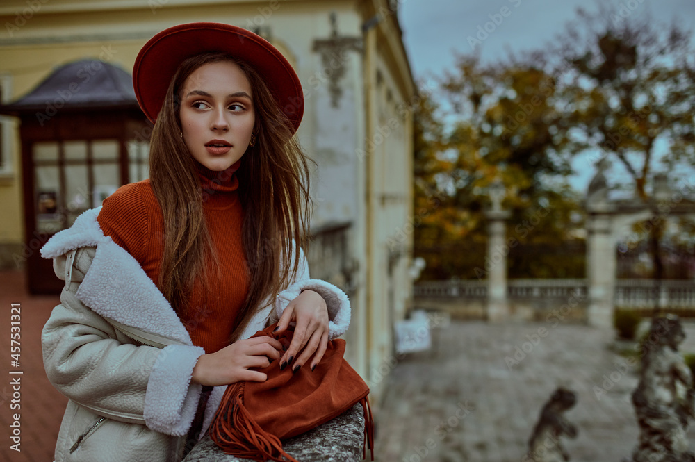 Obraz premium Autumn fashion portrait of elegant woman wearing trendy autumn outfit with orange hat, turtleneck, sheepskin jacket, holding suede bag with fringes, posing in street of European city. Copy space